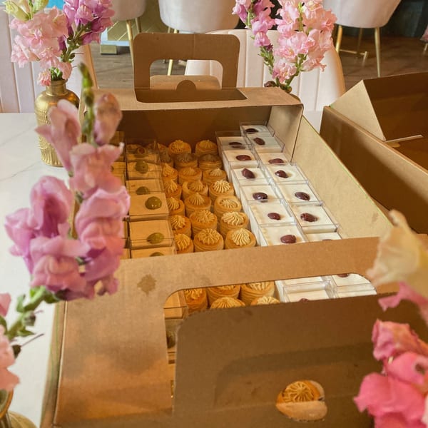 Two cardboard boxes filled with small desserts, including pastries and cups with red fruit, on a table with pink flowers.
