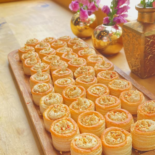 A wooden tray filled with vol-au-vents, each topped with creamy filling and spices, next to golden vases with pink flowers.