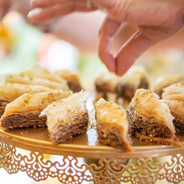 A hand sprinkles nuts over golden baklava pastries arranged on a decorative gold stand.