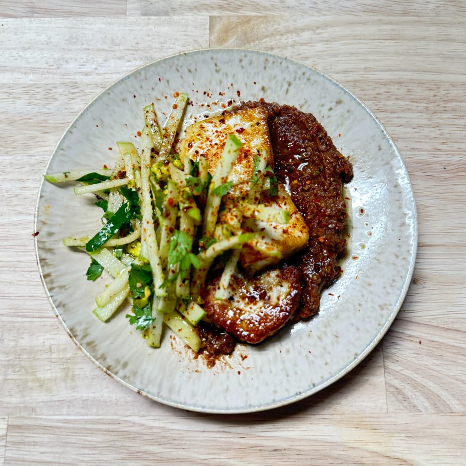 Middle-Eastern dish with seared halloumi, a rich red sauce, and a fresh green salad on a speckled plate.