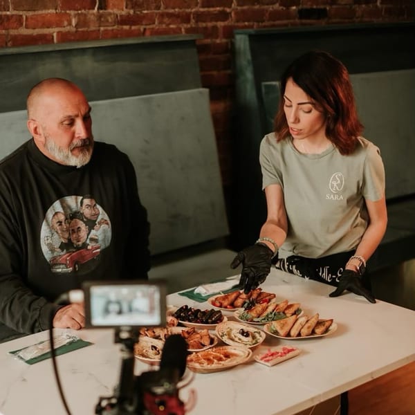 Two people presenting a variety of Middle Eastern dishes, including hummus, dolmas, and pastries.