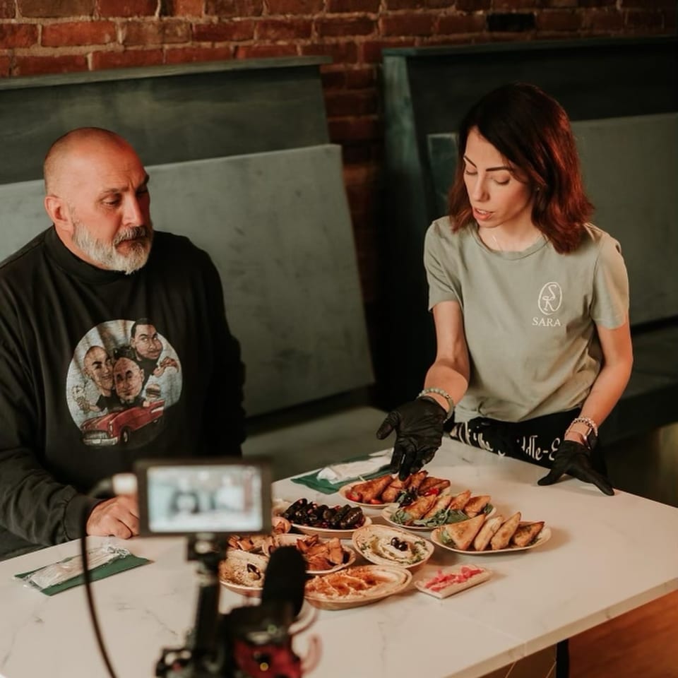 Two people presenting a variety of Middle Eastern dishes, including hummus, dolmas, and pastries.