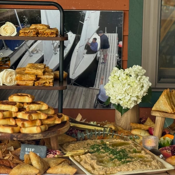 A buffet table with various pastries, dips, and a floral arrangement, against a sailboat backdrop.