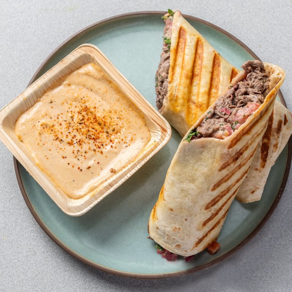 Two grilled Middle Eastern wraps filled with meat and vegetables, served with a side of creamy sauce.