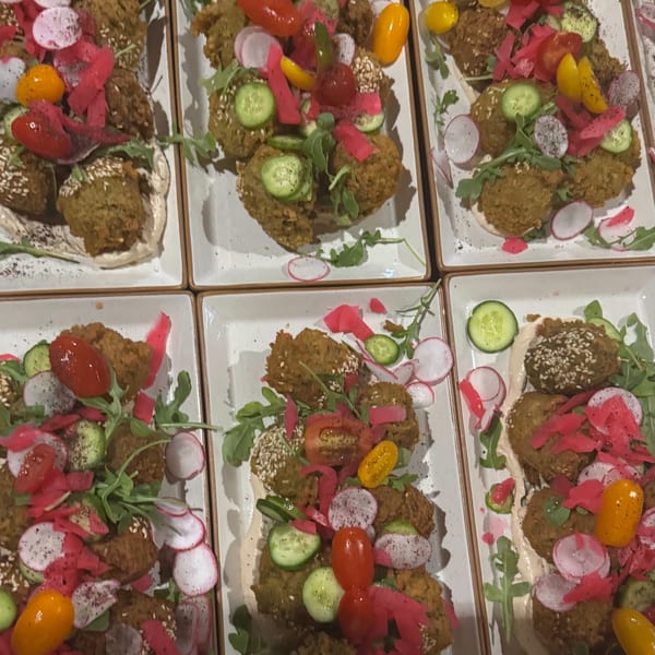 Multiple plates of falafel with hummus, arugula, pickled radishes, cucumbers, and cherry tomatoes.
