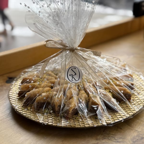 A golden tray of baklava pastries, wrapped in clear, polka-dotted cellophane with a gold ribbon.