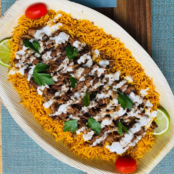 Middle-Eastern rice dish with meat, white sauce, parsley, lime, and tomatoes on a wooden platter.