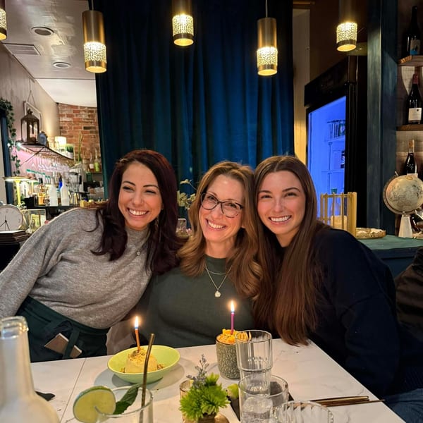 Three smiling women celebrating a birthday with dessert and candles at Sara's Kitchen.