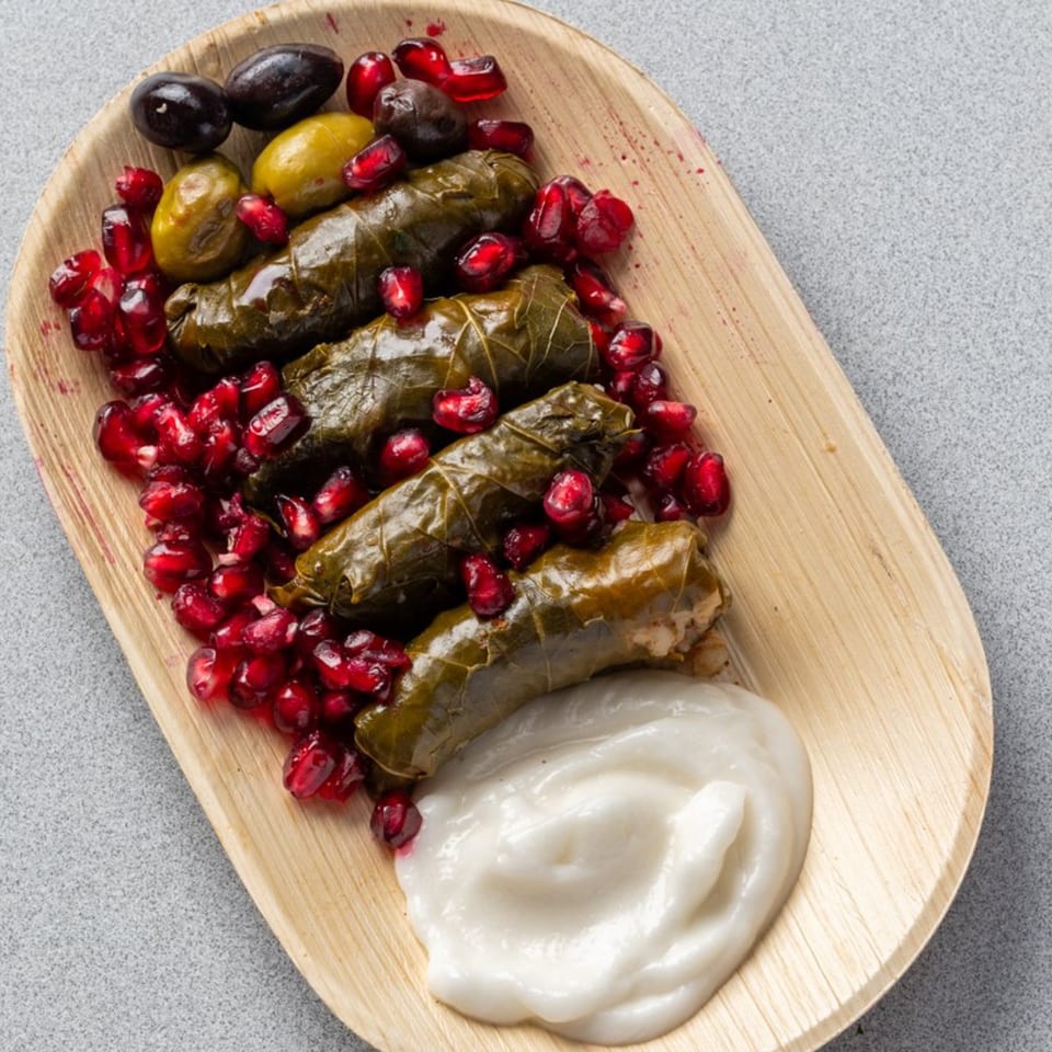 Dolmades with pomegranate seeds, olives, and a white sauce on a wooden plate.