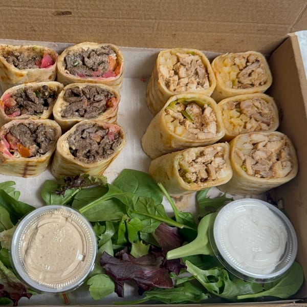 A takeout box filled with beef and chicken shawarma wraps, fresh greens, and two dipping sauces.