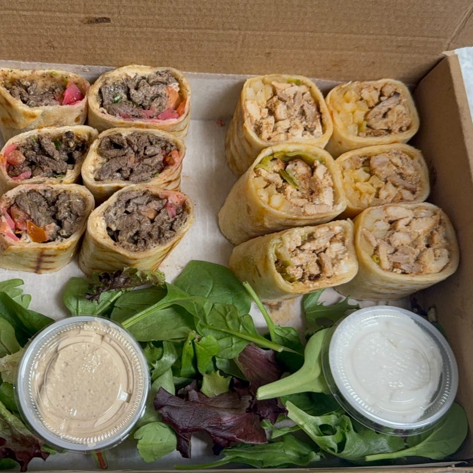 A takeout box filled with beef and chicken shawarma wraps, fresh greens, and two dipping sauces.